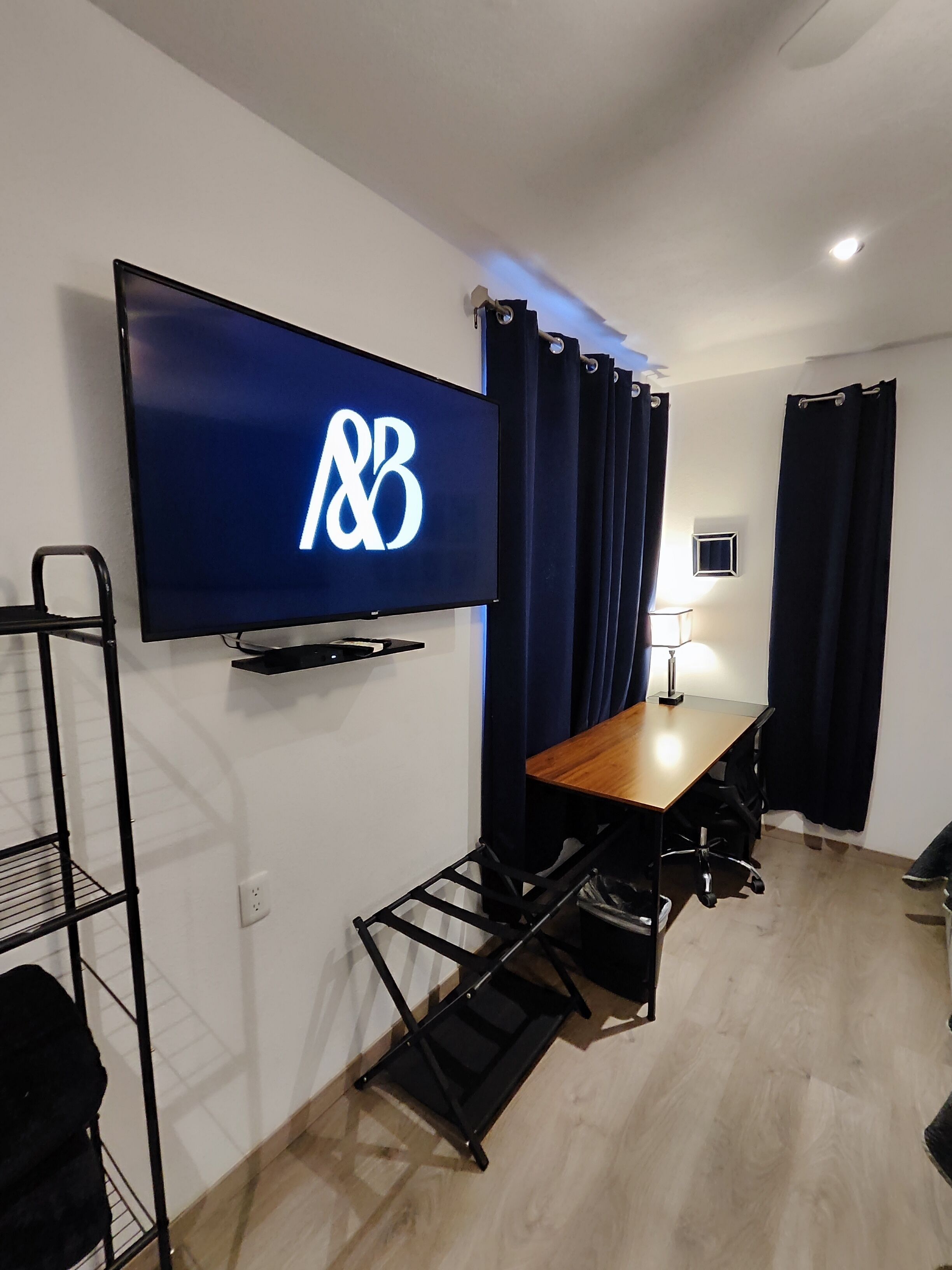 Habitación con una televisión montada que muestra un logotipo, un escritorio con una silla, una lámpara y una ventana con cortinas.