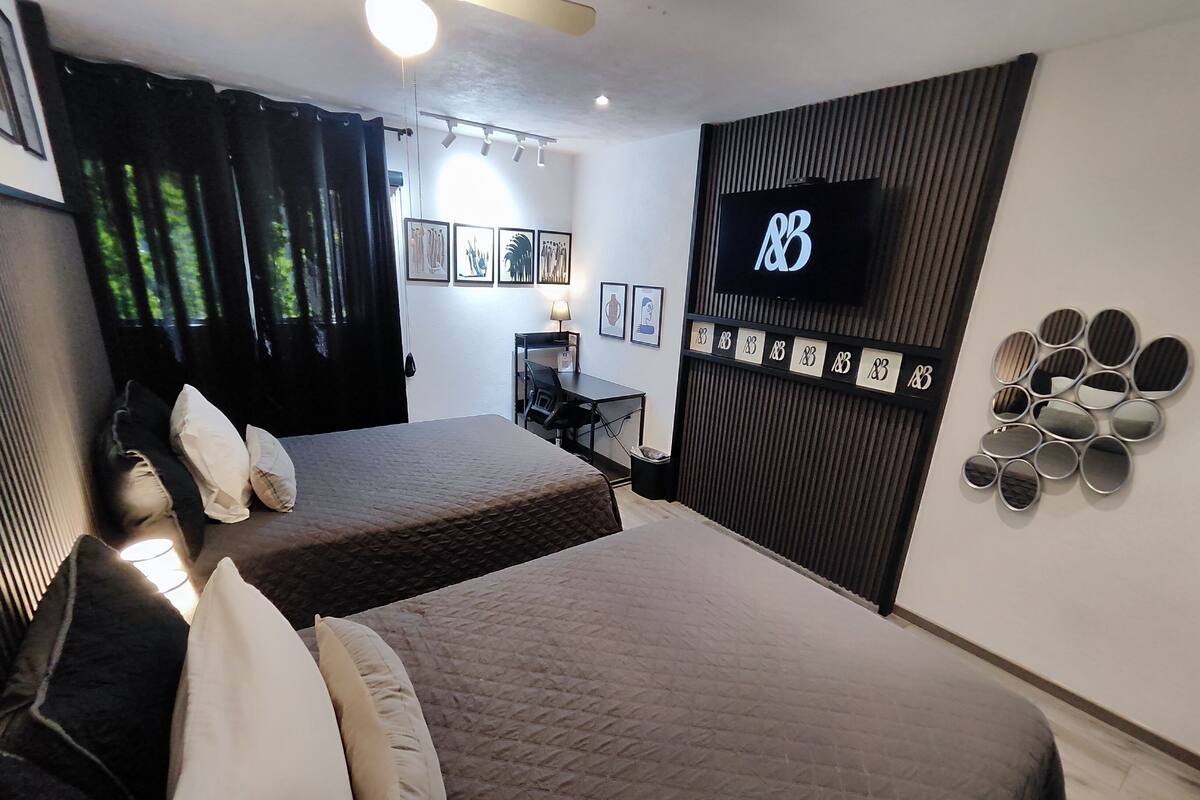 A hotel room with two beds, a desk, a TV, and a wall with framed pictures and a clock.