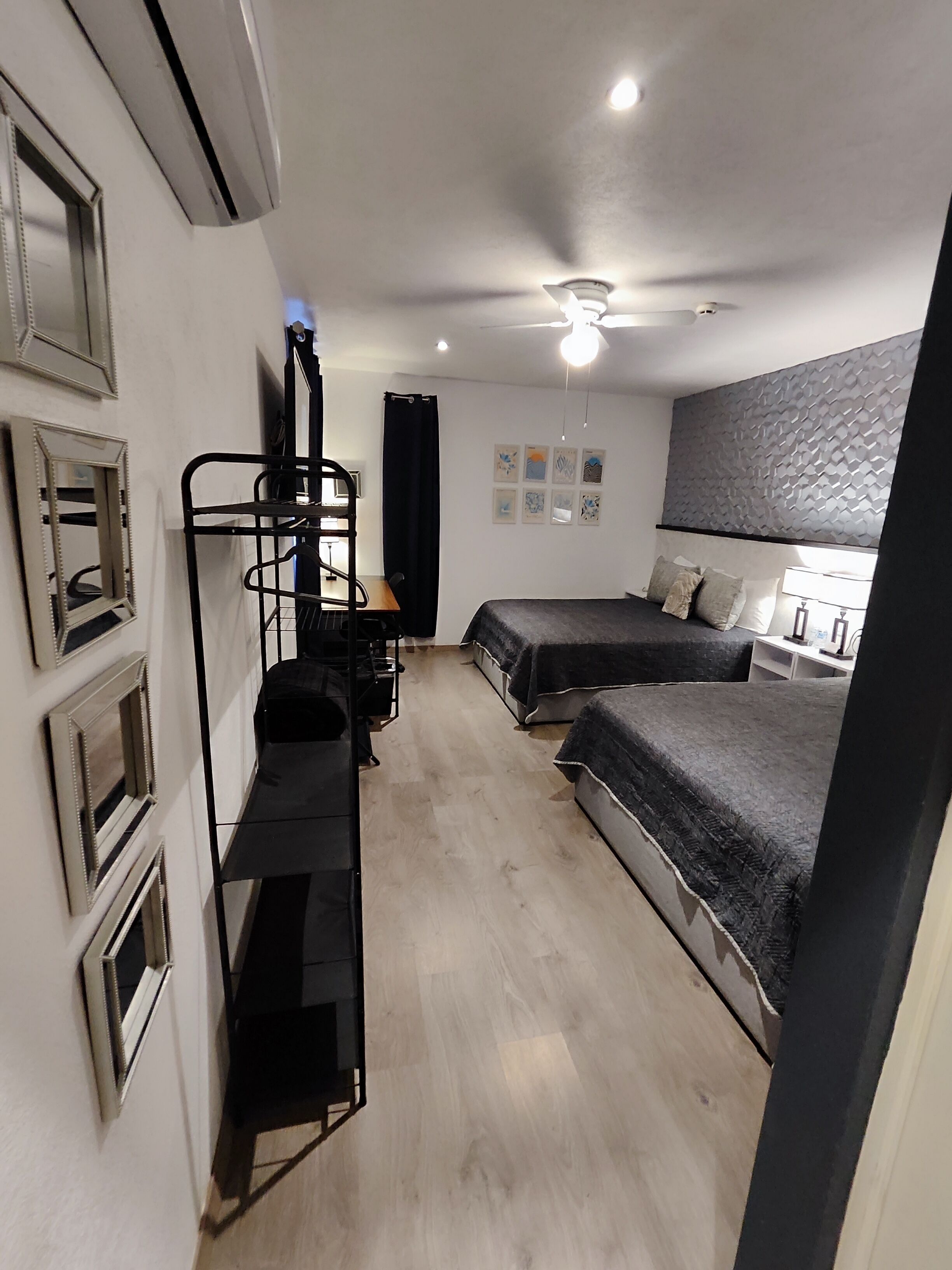 Une chambre de taille modeste, comprenant deux lits, un bureau, une chaise, un ventilateur de plafond et une étagère fixée au mur.