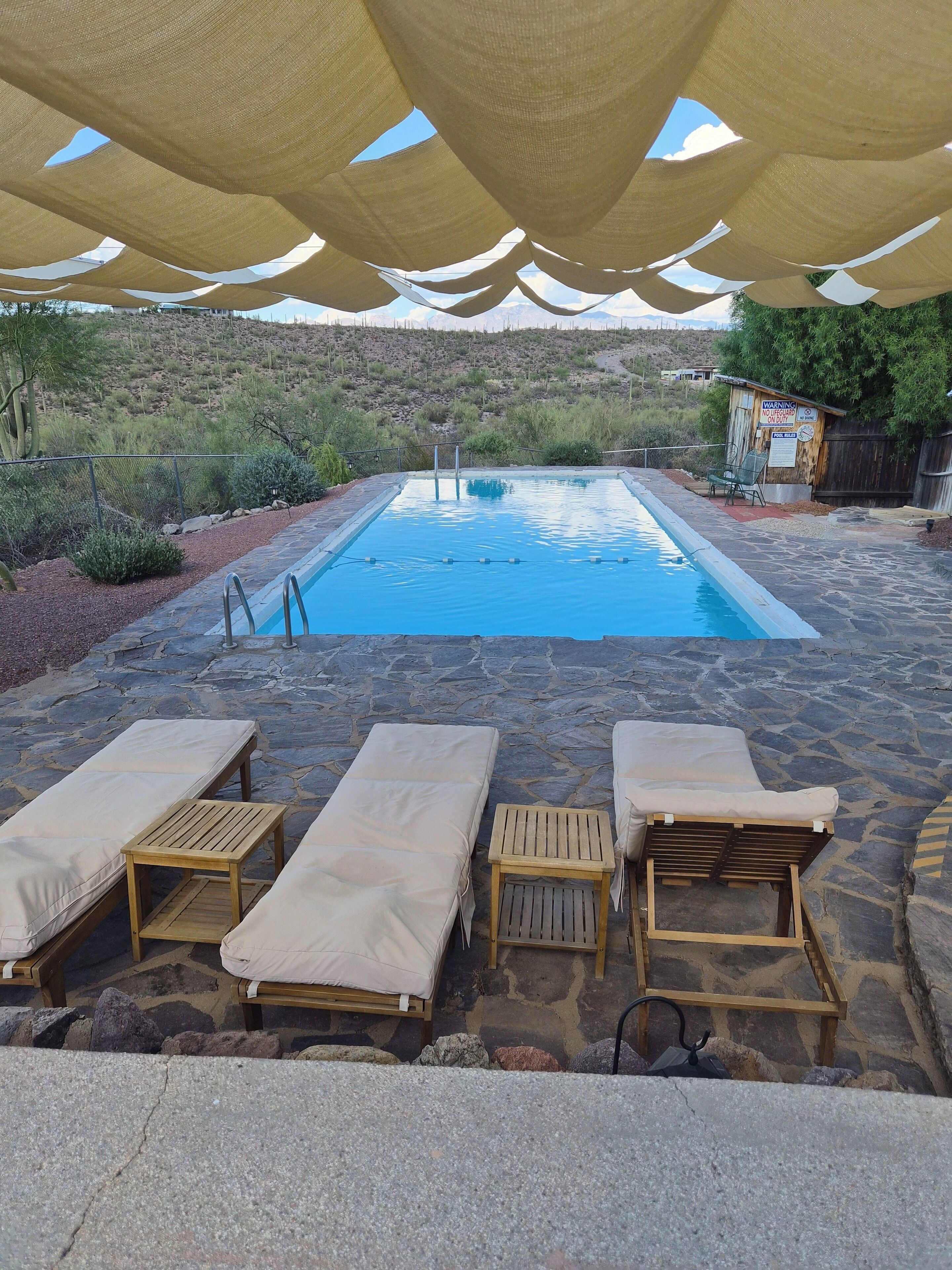 A pool area with lounge chairs and a wooden table under a canopy.