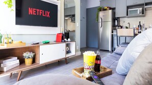 A living room with a TV displaying 'NETFLIX', a popcorn bucket, a remote control, and a bottle of beer on a tray. - Apt piscina e AC com academia e vaga (São Paulo)