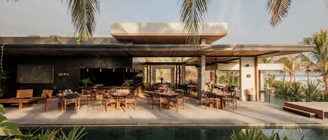 A modern outdoor dining area with a pool, wooden furniture, and tropical plants.