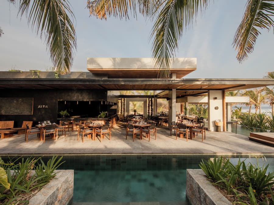 A modern outdoor dining area with a pool, wooden furniture, and tropical plants.