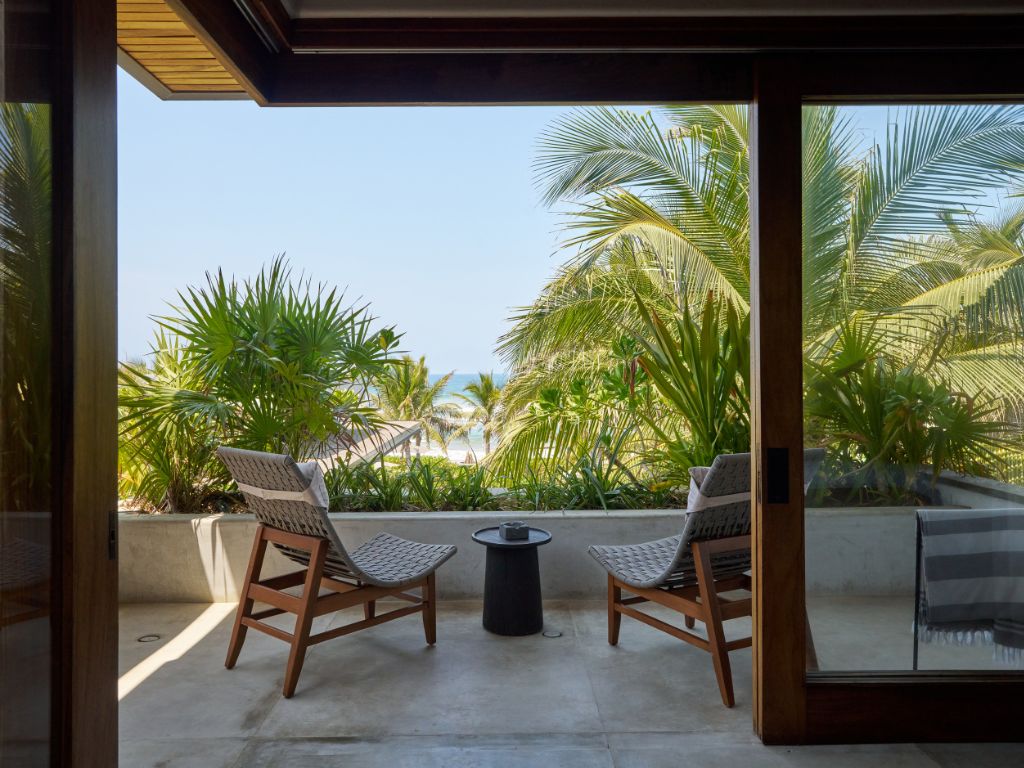 Un balcon avec deux fauteuils en osier et une petite table ronde, donnant sur un jardin tropical avec des palmiers et un ciel dégagé.