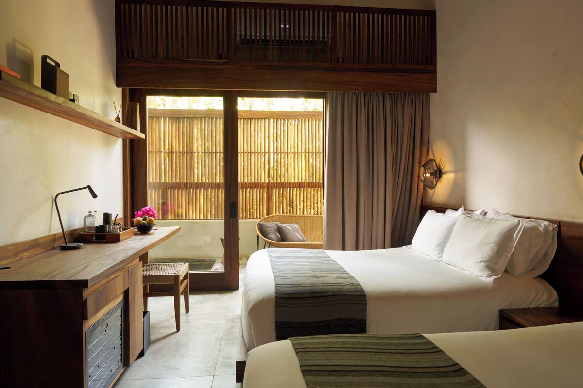 A hotel room with a bed, a desk, a chair, a window with blinds, and a wall-mounted lamp.