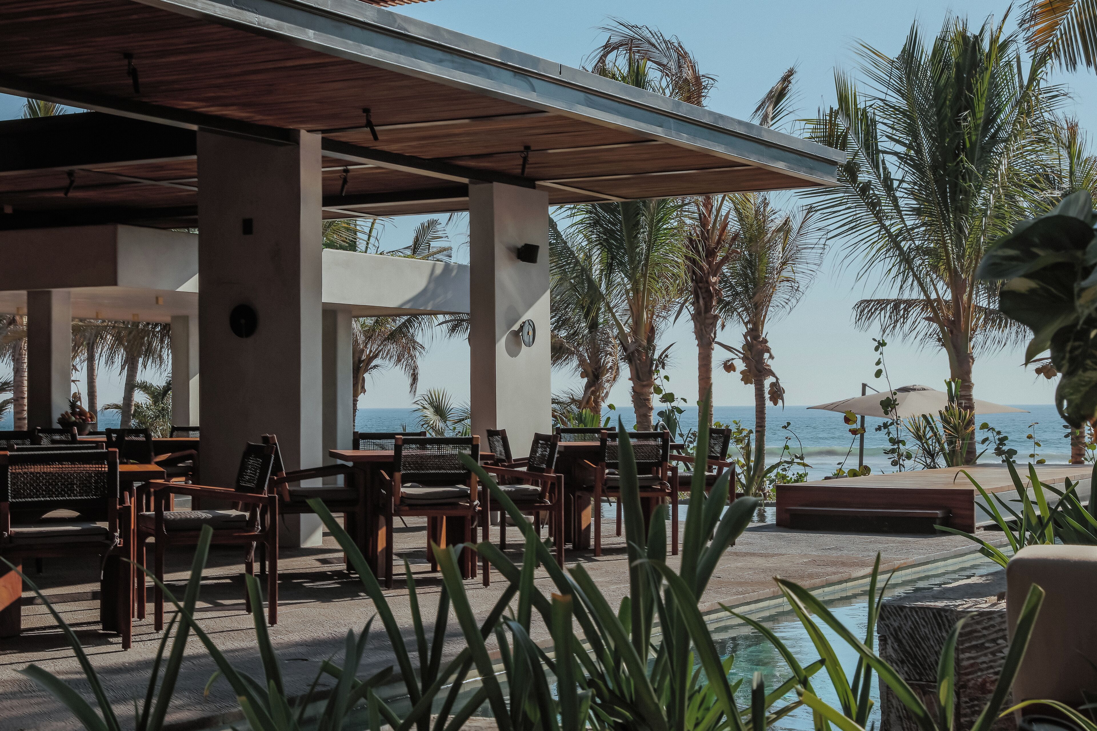 Un área de estar al aire libre con muebles de madera y piscina, rodeada de palmeras y vistas al mar.