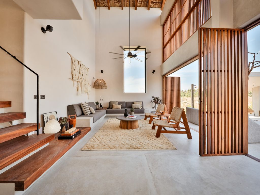 A spacious living area with a sofa, coffee table, and wooden chairs. A staircase is visible on the left, and a large window provides natural light.