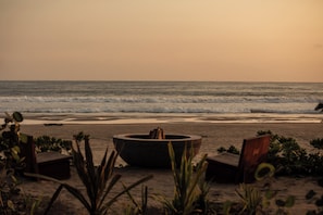 A beach with a fire pit, two wooden chairs, and some plants.