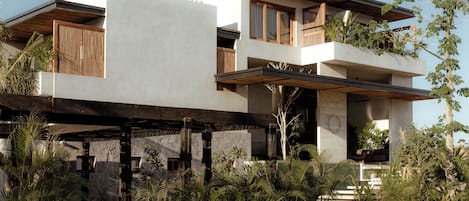 A modern two-story house with a flat roof, large windows, and a covered outdoor area with plants.