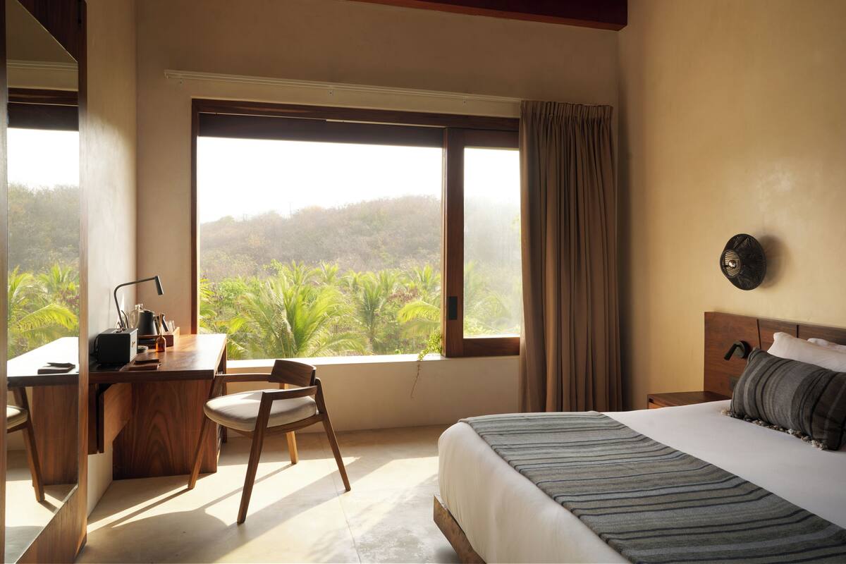 A bedroom with a bed, a desk, a chair, a window with a view of greenery, and a wall-mounted light fixture.
