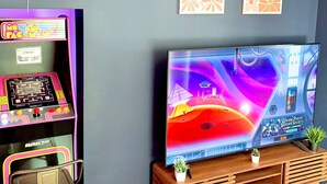 Eine Retro-Spieluhr mit Neonlichtern, ein großer Flachbildfernseher, der ein farbenfrohes Weltraumspiel zeigt, und eine Holz-Unterhaltungsanlage mit einer kleinen Pflanze.