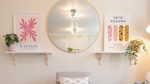Una habitación con un espejo redondo, dos cuadros enmarcados, un sillón gris y una mesita con una planta.