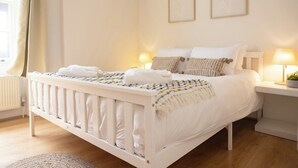 Un dormitorio con una cuna blanca, una cama con ropa de cama blanca, dos lámparas de noche y dos cuadros enmarcados.