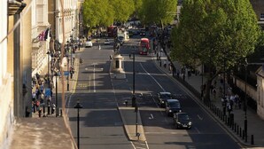 Vista de una calle con edificios históricos, incluyendo una imponente torre de reloj, y una clara vista del Palacio de Westminster al fondo.