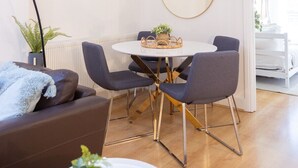 Una sala moderna con un sofá marrón, una mesa de comedor redonda con cuatro sillas y un espejo grande en la pared. Hay obras de arte enmarcadas sobre el espejo y una planta en maceta en el suelo.