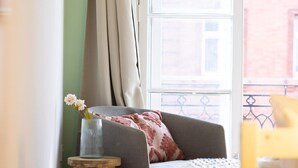 Un dormitorio acogedor con cama, una silla, una mesita con un jarrón de flores y una ventana con cortinas.