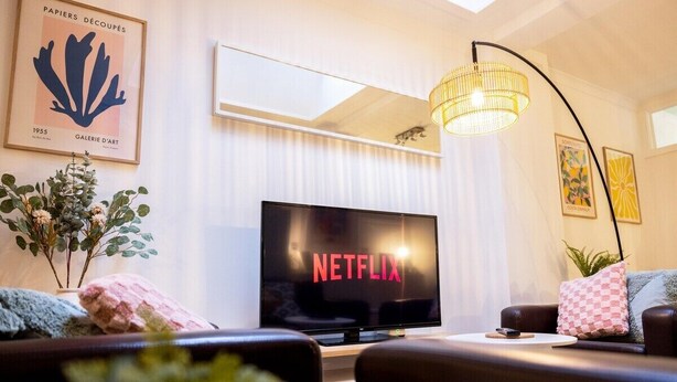 Una sala de estar con televisor de pantalla plana mostrando 'NETFLIX', una lámpara de araña moderna y obras de arte enmarcadas en las paredes.