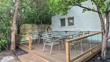 Eine Terrasse im Freien mit Tisch und Stühlen, umgeben von Bäumen und einem weißen Gebäude.