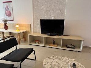 Een woonkamer met een flatscreen televisie op een houten standaard, een zwarte stoel, een ronde marmeren salontafel en een klein boekenrek met boeken.