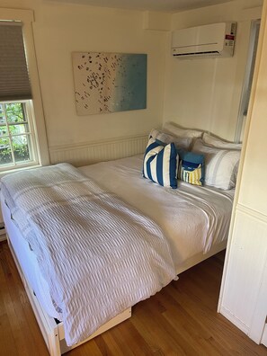 Una cama bien hecha con un almohadón a rayas, una ventana con persianas y un aire acondicionado de pared.