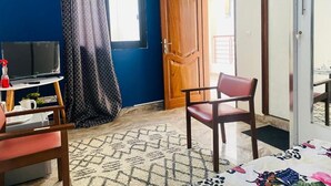 A room with a blue wall, a wooden door, a TV on a stand, a chair, and a patterned rug. - RESIDENCE EL HADJI (Dakar)