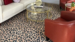 A living room with a sofa, red armchair, glass coffee table, leopard print carpet, framed artwork, and a television.