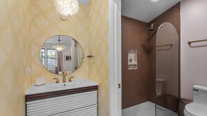 A bathroom with a white sink, gold faucets, a round mirror, and a shower enclosure.