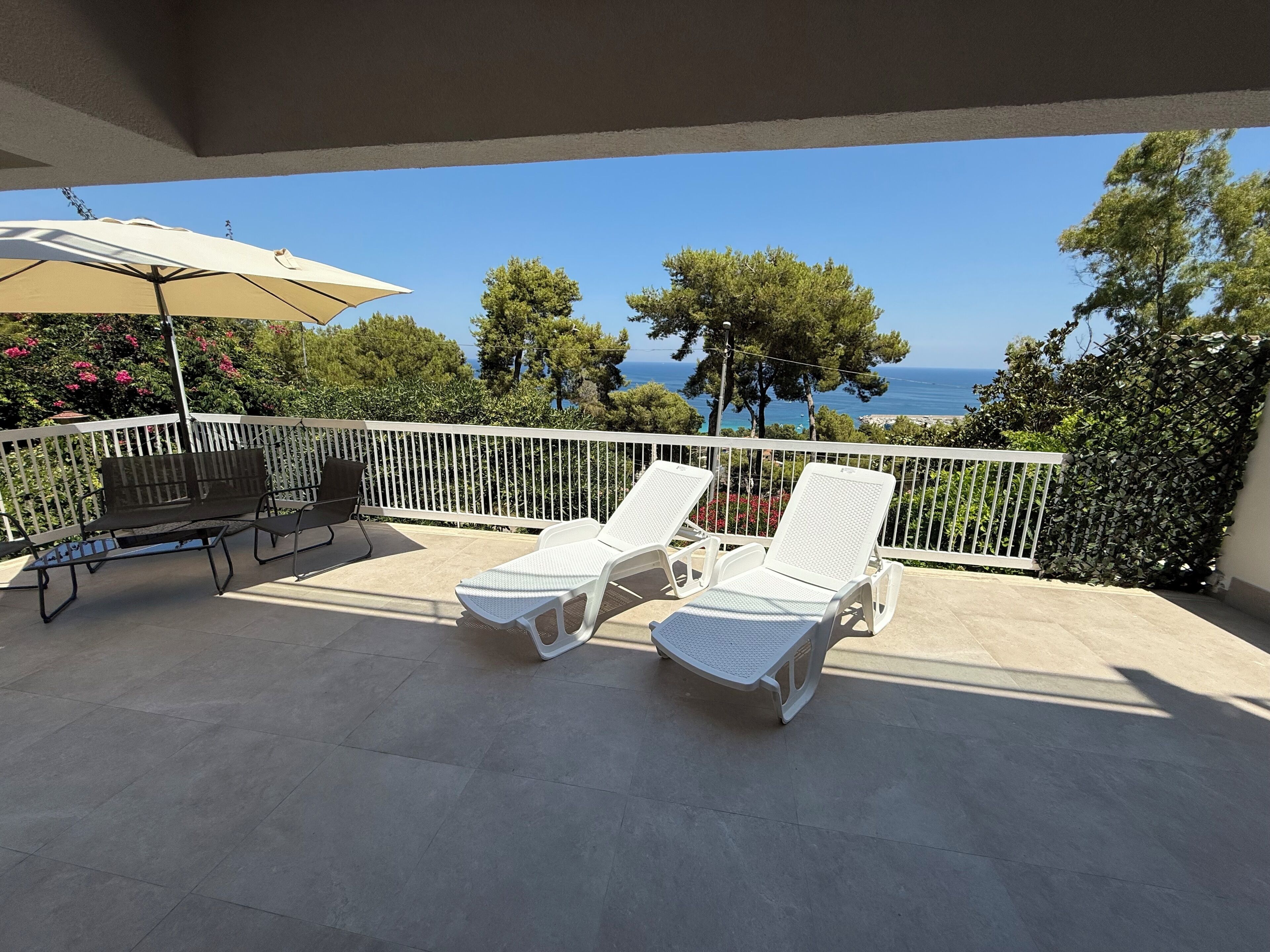 A balcony with two white lounge chairs, a table with a chair, and a beige umbrella.