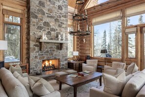 A log cabin interior with a stone fireplace, wooden furniture, and large windows offering a view of a snowy forest.