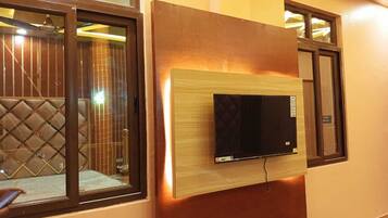 A modern living room with a flat-screen TV mounted on a wooden panel, large windows with dark frames, and a cushioned seating area.