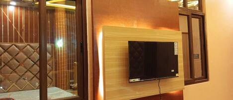 A modern living room with a flat-screen TV mounted on a wooden panel, large windows with dark frames, and a cushioned seating area.