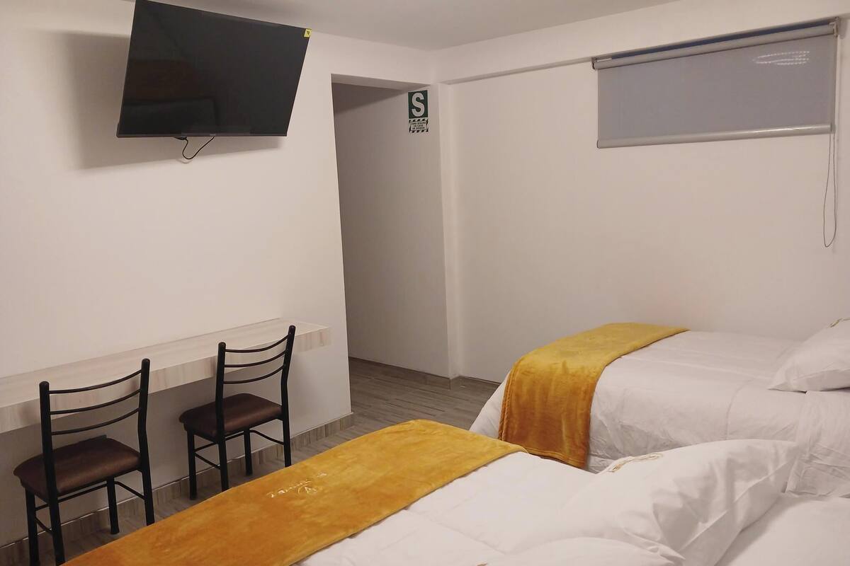 A hotel room with two beds, a TV, a desk with chairs, and a window with a blind.