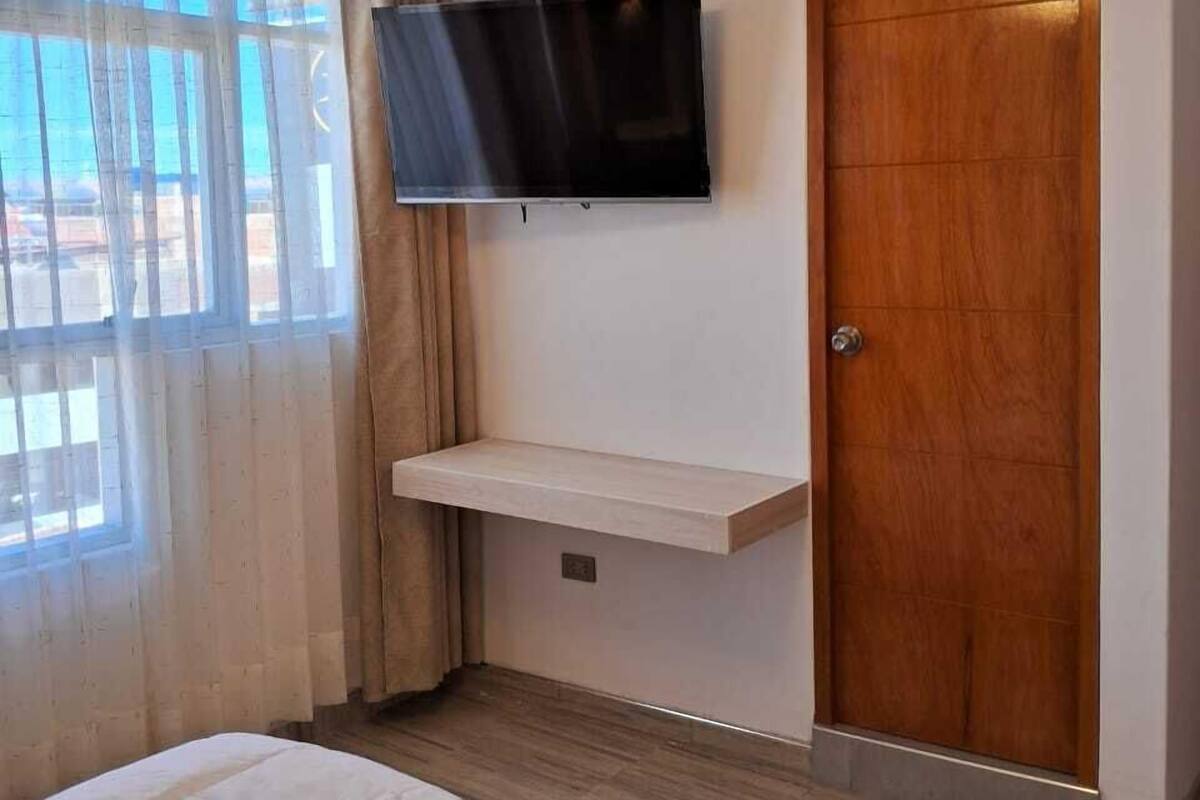A hotel room with a bed, a mounted TV, a wooden door, and a window with curtains.