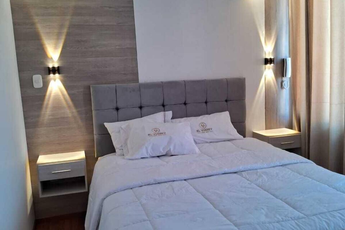 A modern bedroom with a large bed, two bedside tables with integrated lighting, a wooden accent wall, and a neutral color scheme.