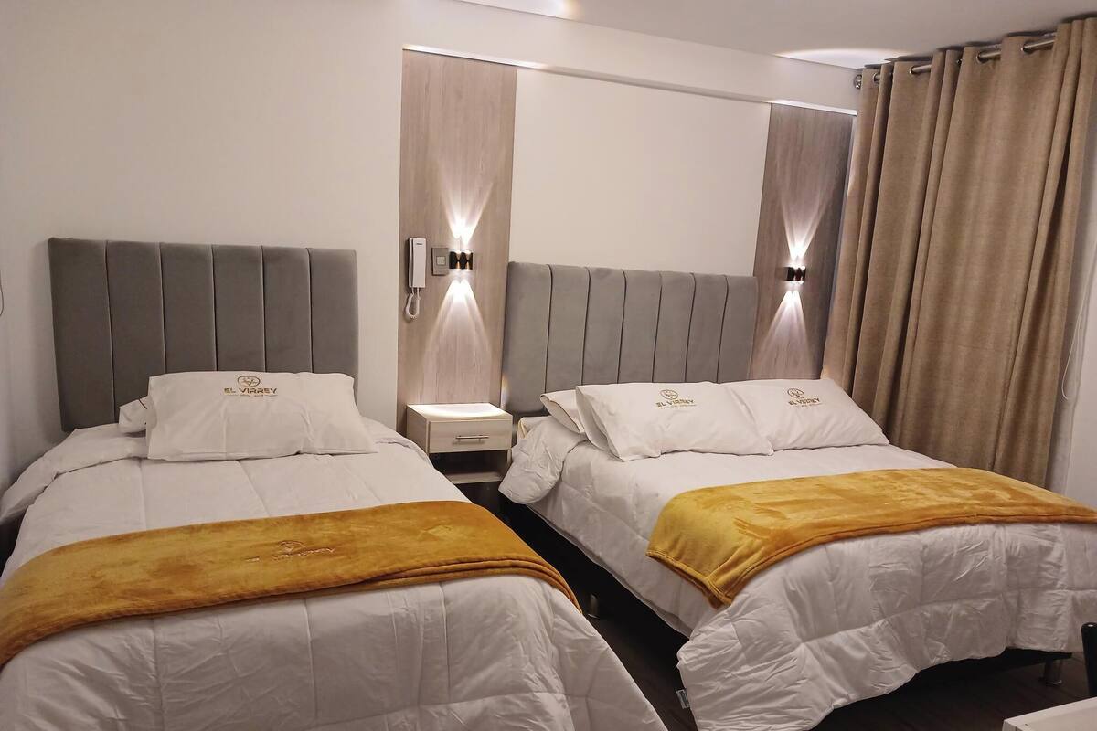 A hotel room with two beds, each with a headboard and a bedside table. There is a wall-mounted phone and a light fixture above the beds.