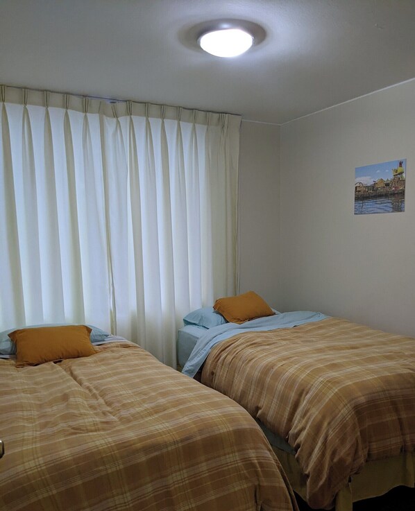 Habitación con dos camas, cada una con un edredón de diferente color, y una ventana con cortinas blancas.