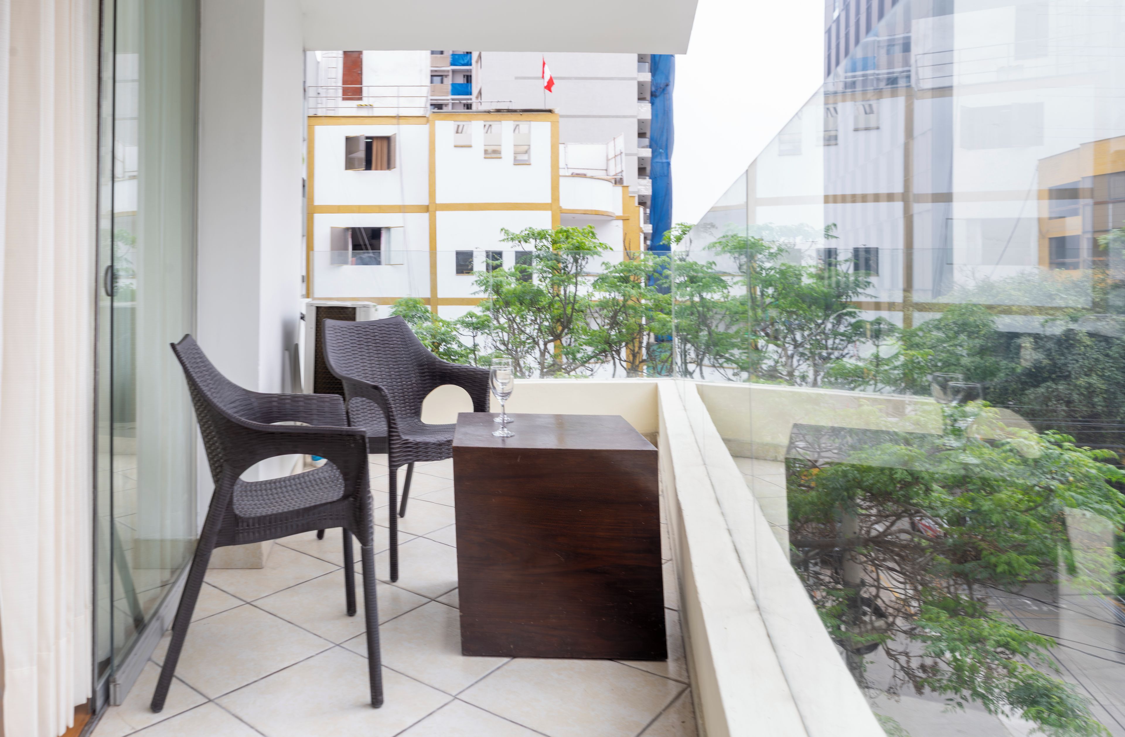 Un balcone con due poltrone in vimini e un tavolino, affacciato su un panorama urbano con edifici e aree verdi.