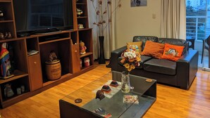 Ett vardagsrum med en tv, trähyllor, ett soffbord i glas och en soffa med orange kuddar.