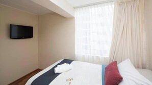 Ett hotellrum med en säng, en platt-TV som är monterad på väggen, ett fönster med tunna, genomskinliga gardiner och utsikt över stadsbilden.