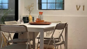 Un comedor con una mesa blanca y cuatro sillas, un jarrón con flores secas y una ventana con diseño de cuadrícula.