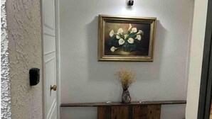 Un pasillo con una puerta blanca, un aplique en la pared, un cuadro enmarcado con flores y un pequeño florero con flores secas sobre una consola de madera.