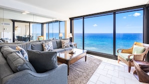 A spacious living room with a large window overlooking the ocean, a sofa set, a coffee table, and a wicker chair.