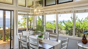 Un espace repas comprenant une table et des chaises, de grandes fenêtres offrant une vue sur la verdure et l’océan, ainsi qu’un luminaire suspendu.