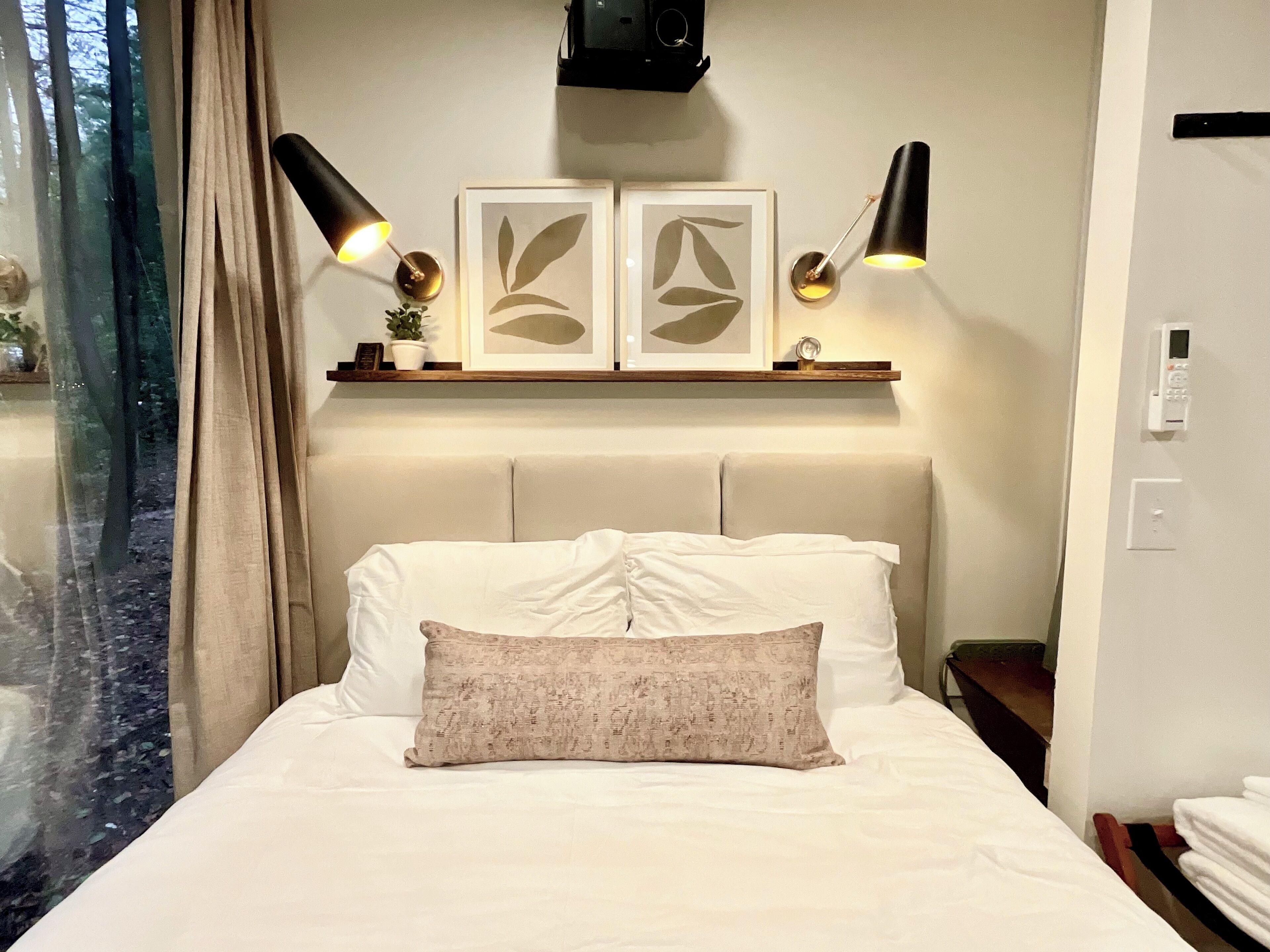 A hotel room with a bed, two wall-mounted lamps, a shelf with framed artwork, and a window with curtains.