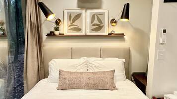 A hotel room with a bed, two wall-mounted lamps, a shelf with framed artwork, and a window with curtains.