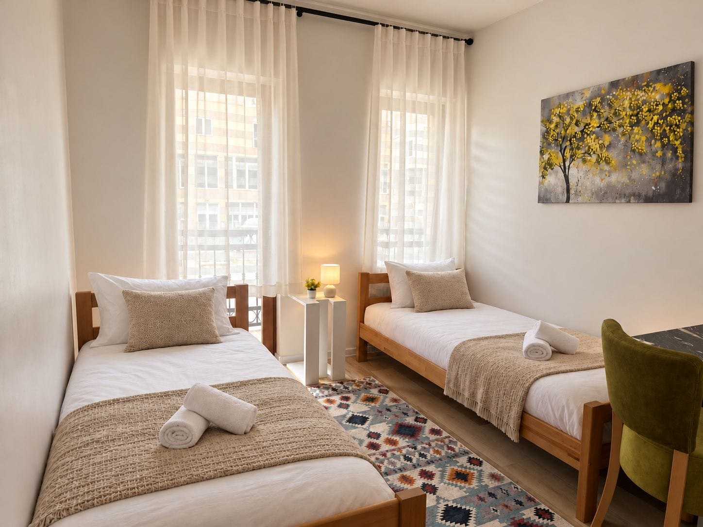 A hotel room with two single beds, a small table, a chair, a painting on the wall, and a patterned rug.