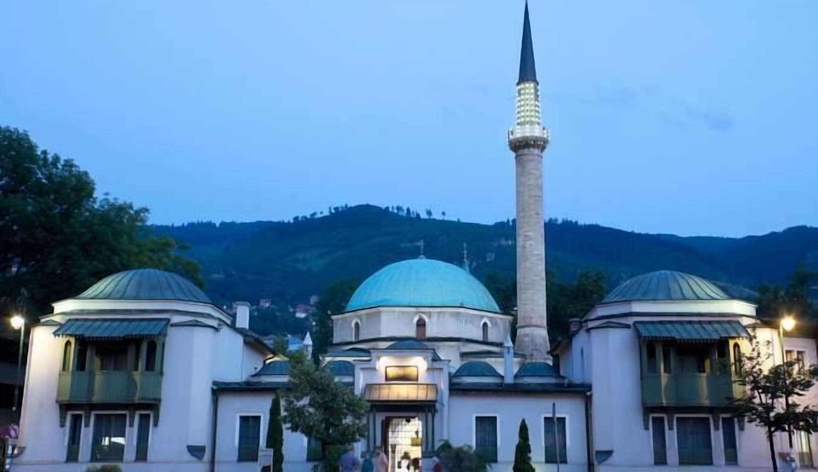 A mosque with two domes and a tall minaret, surrounded by trees and hills.