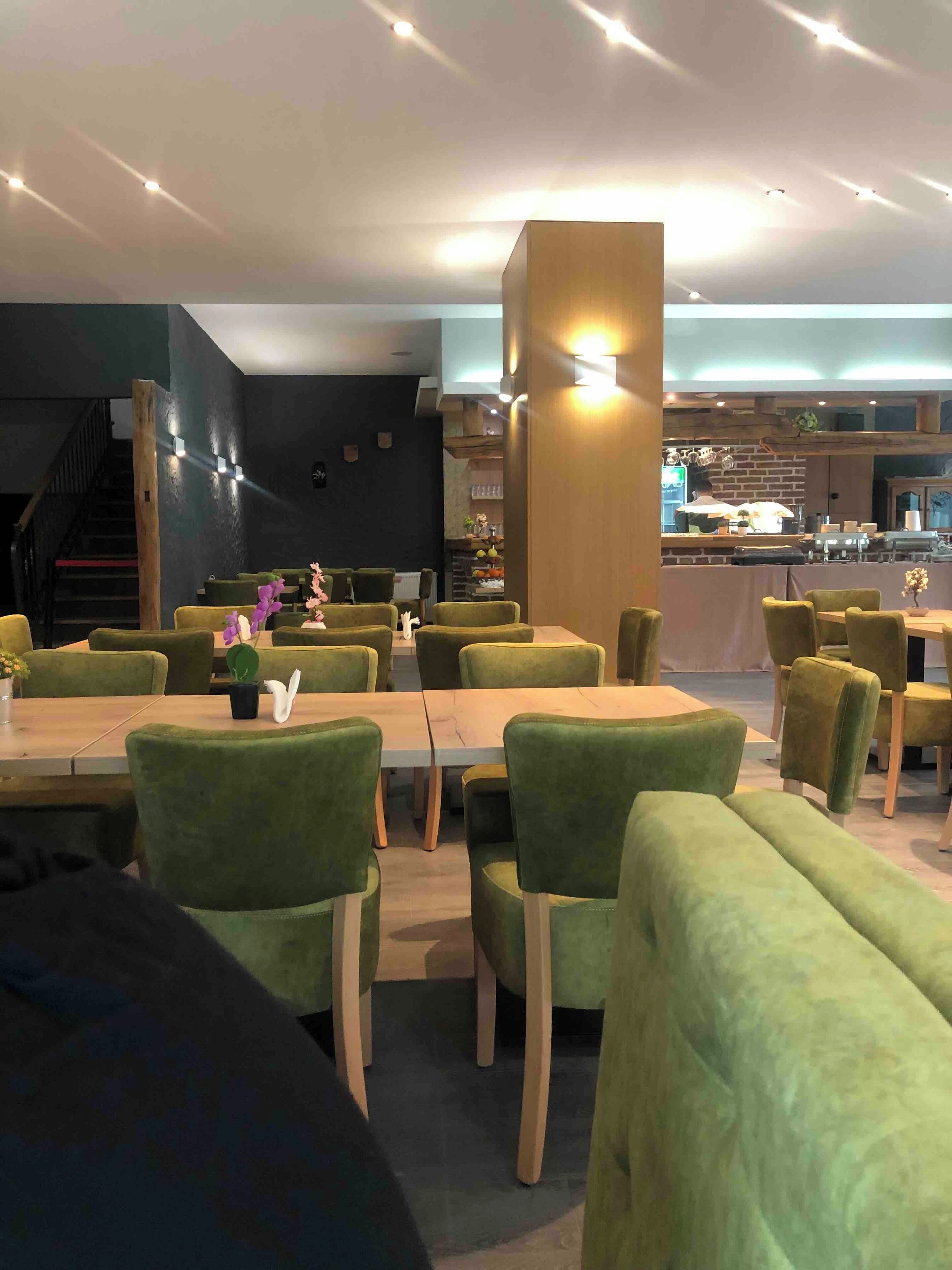 A restaurant with green upholstered chairs and tables, a wooden pillar, and a kitchen area in the background.