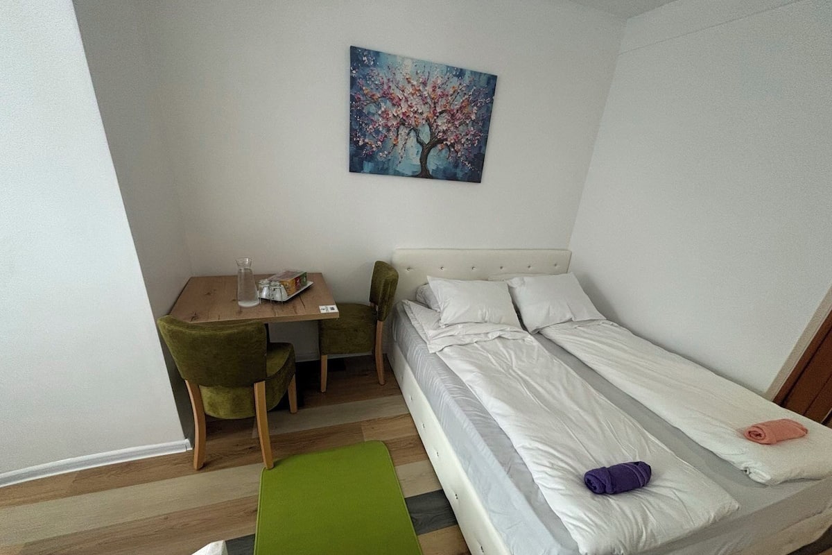 Quarto pequeno e bem arrumado com uma cama, uma cadeira, uma mesa e um quadro na parede.