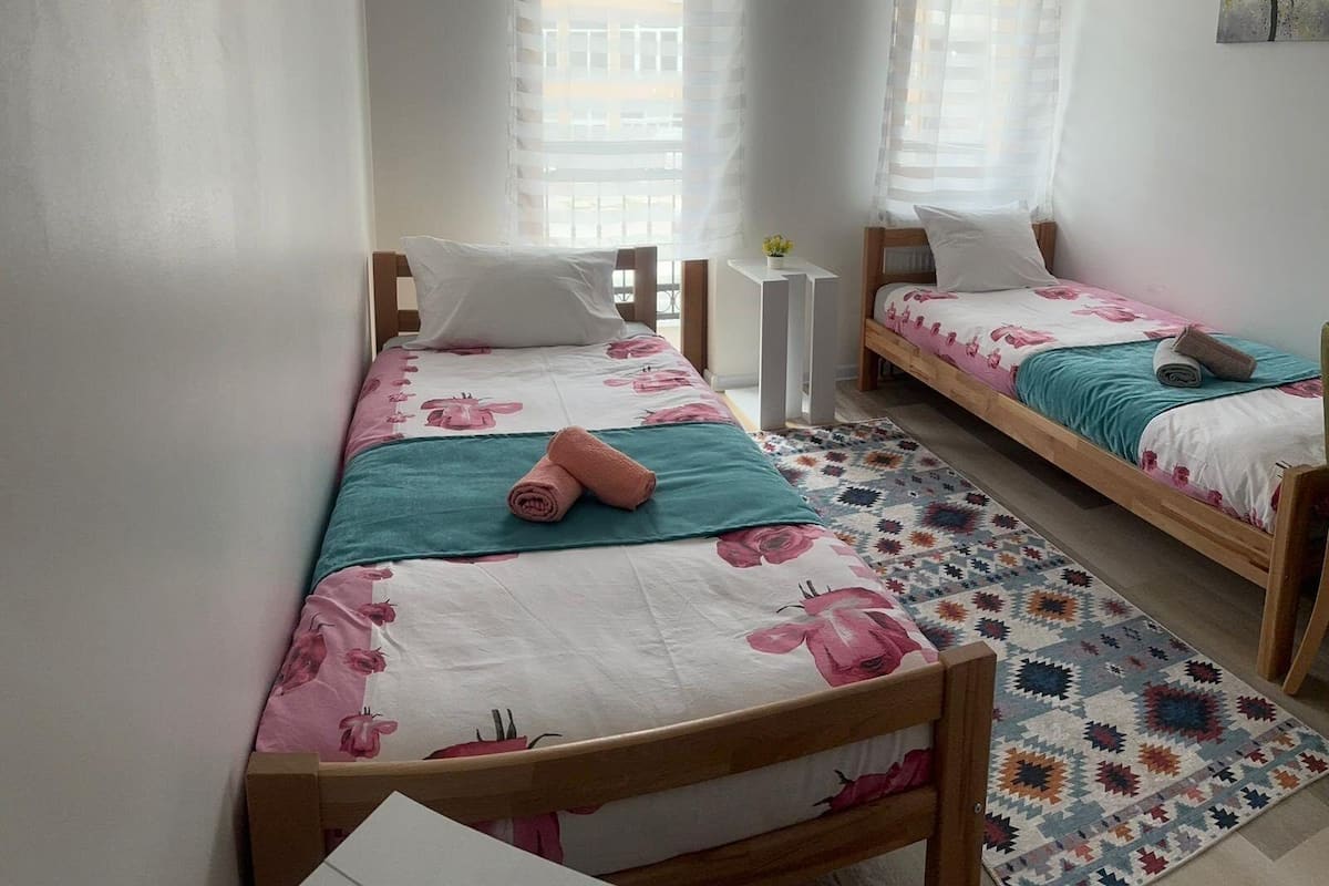 Quarto pequeno e limpo, com duas camas de solteiro, uma mesinha e janela com cortinas leves.
